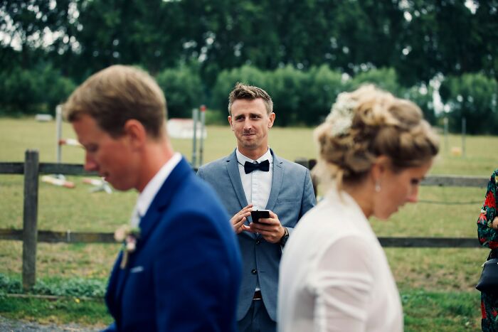 Man in a suit holding a phone at an outdoor wedding, capturing a moment with wedding guests nearby.