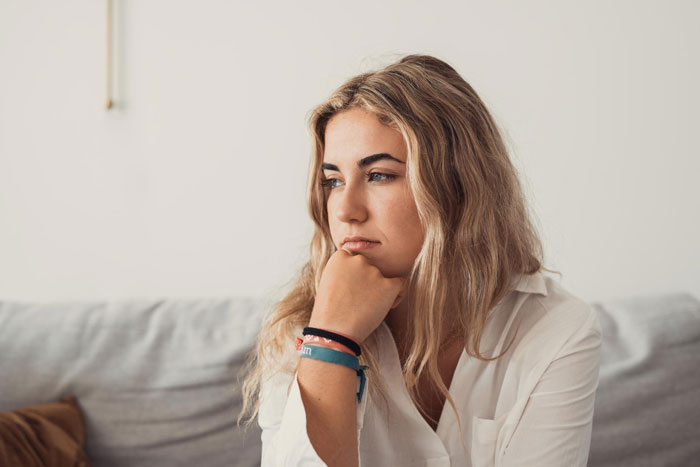 Young woman with blonde hair and white shirt sitting thoughtfully on a couch reflecting on rude MIL behavior issues.