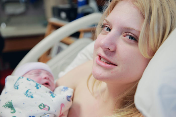 New mother holding newborn baby in hospital bed after giving birth, with overbearing MIL booking photoshoot nearby.