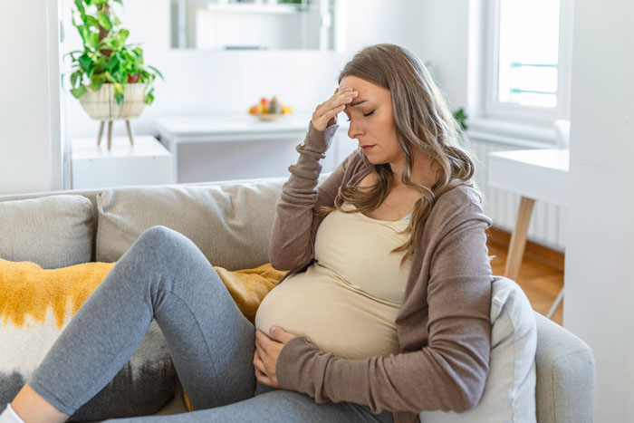 Pregnant woman sitting on couch, looking stressed and holding her belly in a bright living room setting.