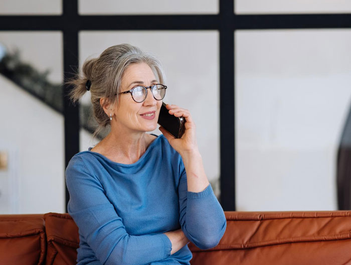 Middle-aged woman wearing glasses sitting on couch, talking on phone, representing DIL receiving mysterious nonstop calls.