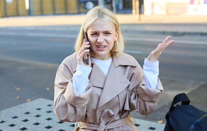 Young woman looking annoyed while talking on phone, upset by nonstop calls from mystery number, confused outdoors.