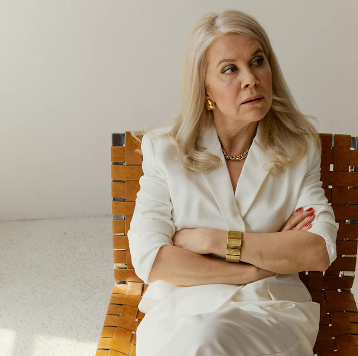 Middle-aged woman in white outfit sitting with crossed arms, appearing upset during wedding attention conflict.