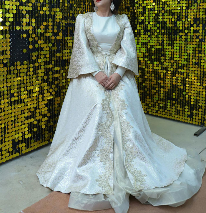 Woman in ornate white wedding dress with gold embroidery standing against a sequin gold background at a wedding event.