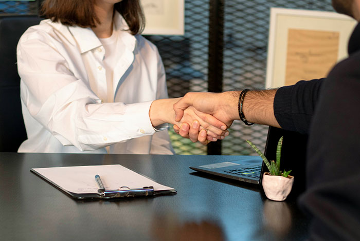 Two people shaking hands across a table with a laptop and clipboard, illustrating social cues and cringe moments.