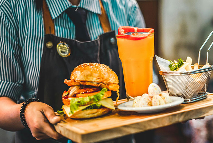 Waiter serving a burger, fries, and a drink on a wooden tray, illustrating social cues and cringe moments in dining.