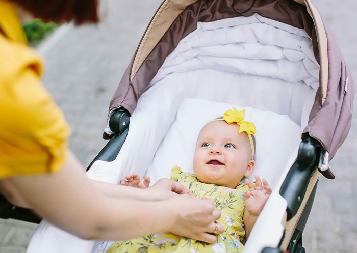 Baby smiling in stroller wearing yellow dress and headband, with adult hands adjusting outfit showing social cues misread.