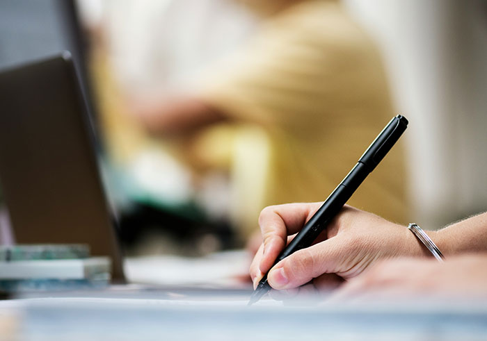 Close-up of a hand holding a pen writing notes on paper with a blurred laptop and person in the background, related to misread social cues.