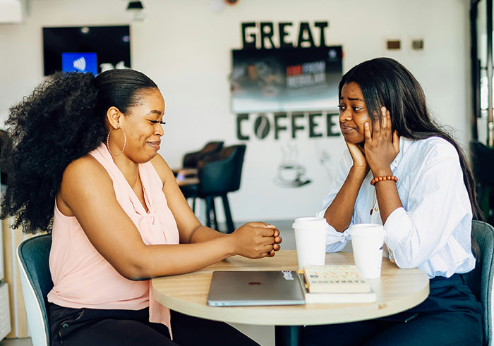 Two women at a coffee shop table, one looking amused and the other reacting awkwardly, showing misread social cues.