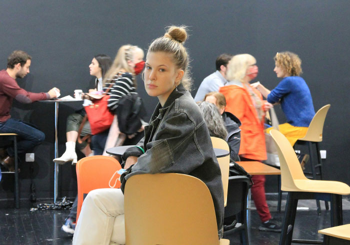 Young woman sitting alone in a public space, appearing thoughtful among busy people, highlighting social cues and cringe moments.
