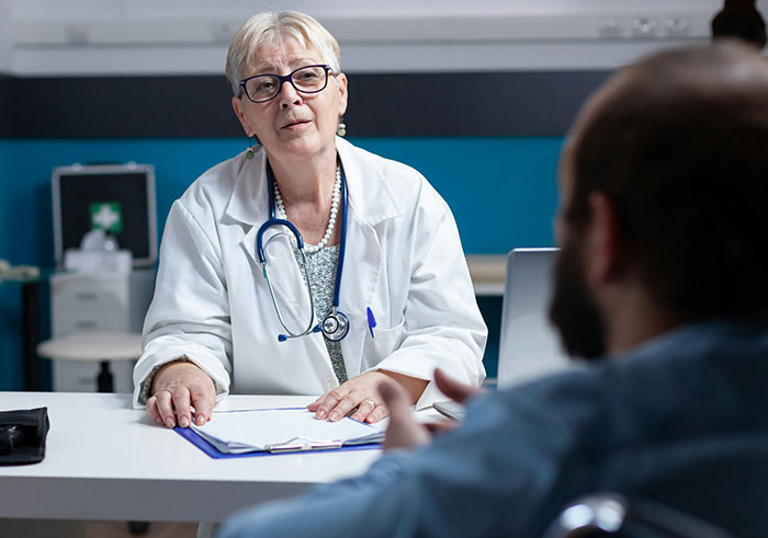 Older female doctor with glasses talks to male patient, highlighting moments of misread social cues and cringe interactions.