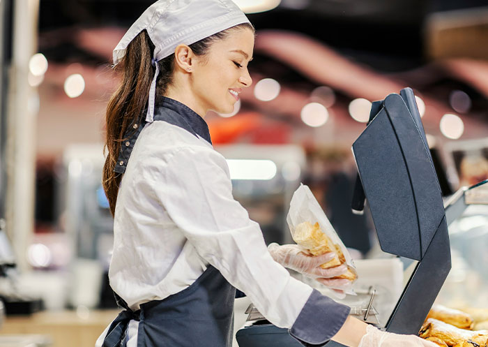 Smiling food service worker wearing uniform and gloves, handling packaged food while operating a cash register in a busy setting.