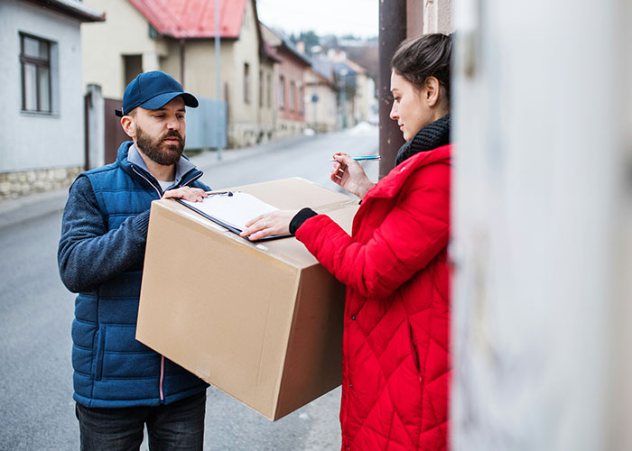 Delivery man holding a large box while woman in red coat awkwardly signs clipboard on a quiet residential street.