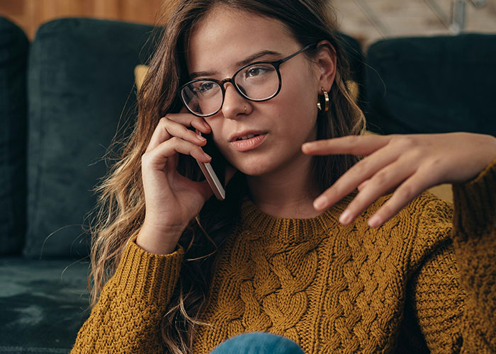 Young woman in glasses on phone showing a socially awkward gesture while sitting on a couch in a cozy sweater