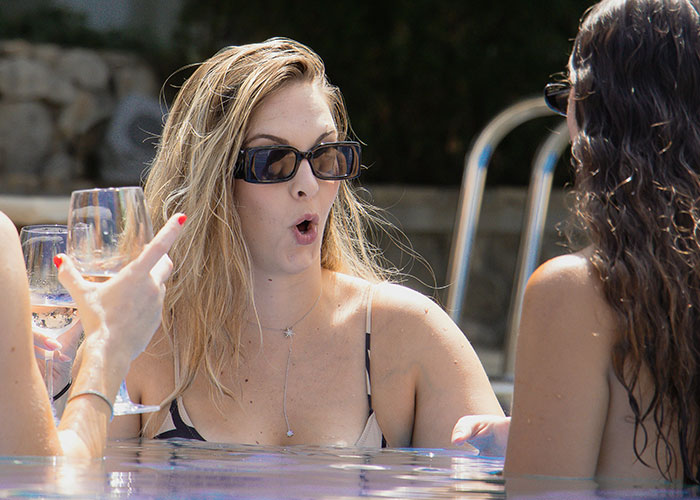 Three women in sunglasses experiencing socially awkward situations while talking and drinking in a pool on a sunny day.