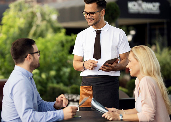 Waiter with a digital tablet serving a couple at an outdoor restaurant, illustrating socially awkward situations.