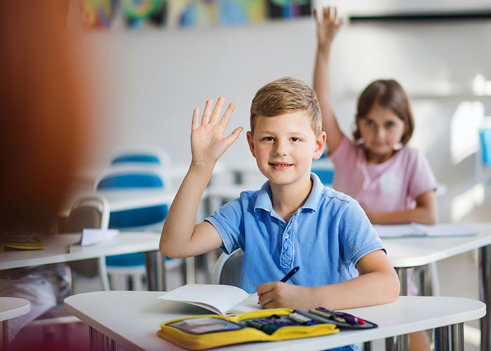 Two children in a classroom raising hands, capturing a socially awkward situation with shy expressions and hesitant smiles.