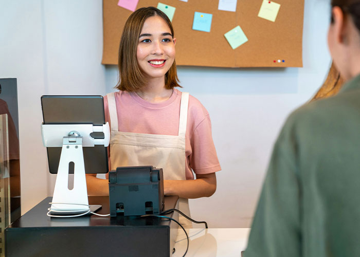 Young female cashier smiling awkwardly while interacting with a customer at a modern point of sale counter.