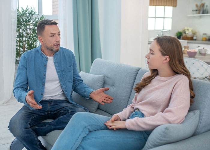 Man and teenage daughter having a serious conversation on a couch, reflecting struggles of mom admitting favoritism between daughters.