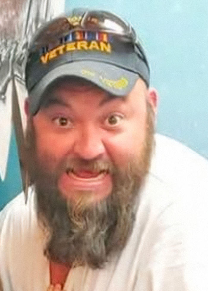 Bearded man wearing a veteran cap with sunglasses, expressing a surprised or intense emotion indoors.
