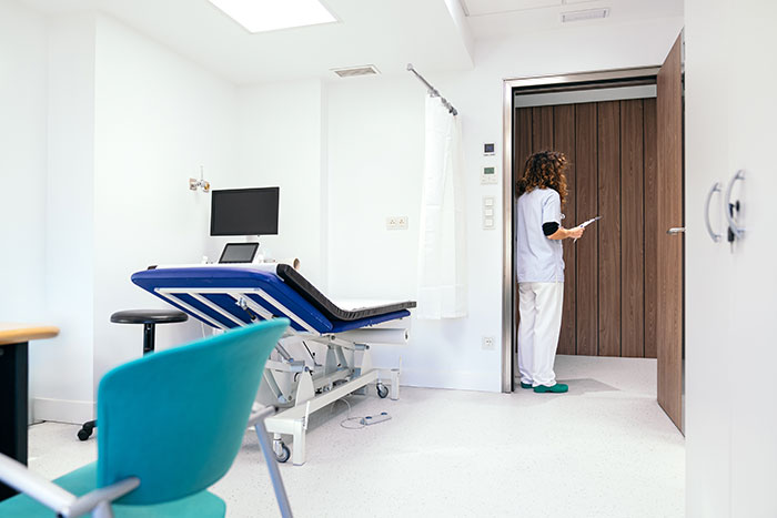 Doctor standing in a medical office with an exam table and computer, representing self-diagnosed patients and doctors.