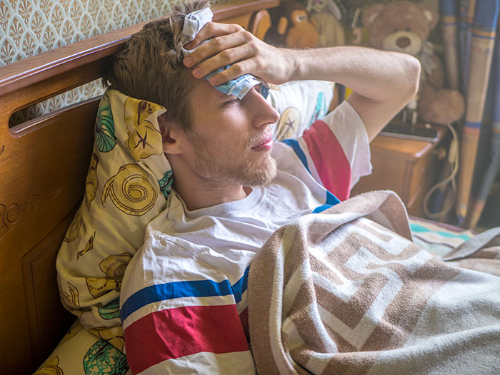 Young man lying in bed holding ice pack to forehead, illustrating self-diagnosed illness and medical correction.