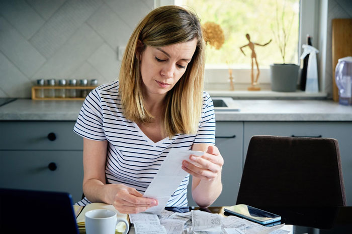 Woman venting about useless bro, looking frustrated while reviewing receipts at kitchen table with phone and laptop nearby