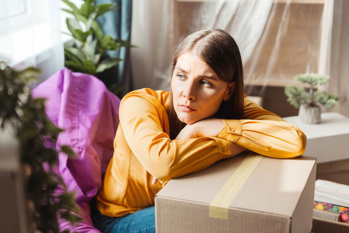 Young woman in a yellow shirt looking upset while leaning on a cardboard box, venting about her useless brother.