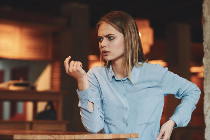 Young woman in a blue shirt looking curious while overhearing multilingual gossip in a cozy indoor setting.