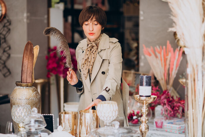 A multilingual woman in a beige coat holding a feather, surrounded by decorative items in an elegant setting.