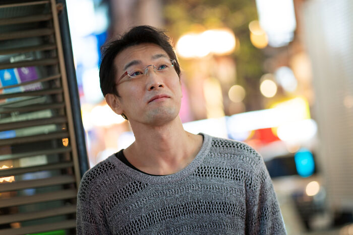 Asian man wearing glasses and a gray sweater, thoughtfully observing people gossiping in secret multilingual languages.