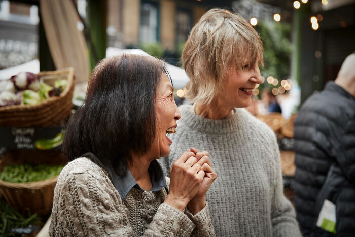 Two multilingual women laughing and catching folks gossiping in secret languages at an outdoor market.