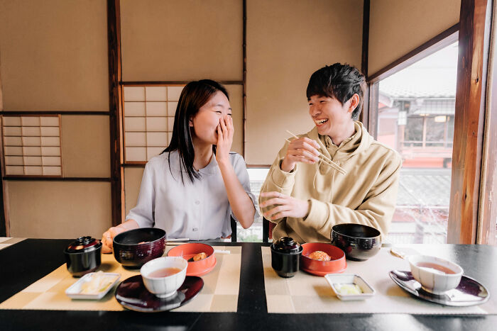 Two multilingual friends laughing and catching gossip while enjoying a traditional meal in a cozy setting.