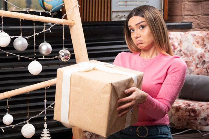 Woman in pink sweater holding a large wrapped gift with a disappointed expression about mums bad birthday presents.