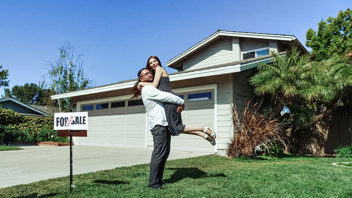 Couple embracing in front of a big home with a for sale sign, highlighting owning a big home with no kids.