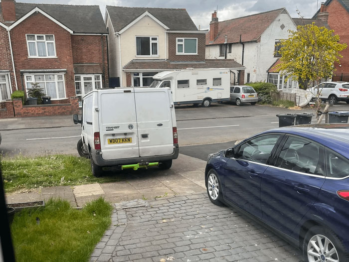 White van blocking neighbor's driveway in a residential area with cars parked nearby on a cloudy day.