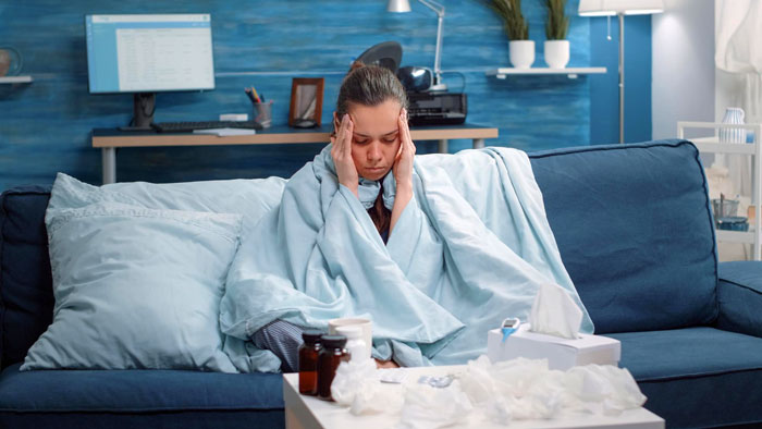 Sick woman wrapped in blanket on couch experiencing headache, surrounded by medicine and tissues, suggesting medical leave concerns.