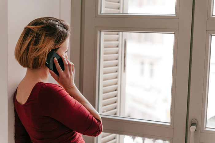 Woman in red shirt standing by window, speaking on phone, depicting neighbor conflict and potential fraud reporting issue.