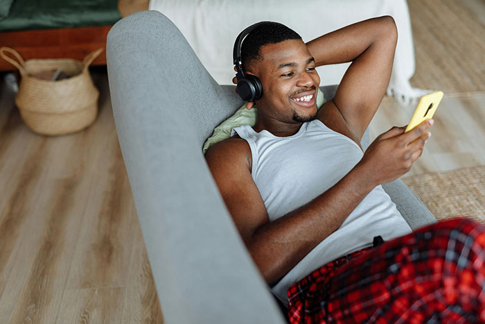 Young man relaxing on couch with headphones, smiling while using phone, illustrating red flags in relationships concept.