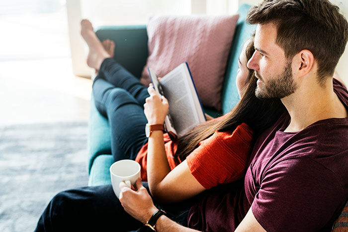 Couple relaxing on a couch, woman reading and man holding a coffee mug, highlighting relationship red flags awareness.