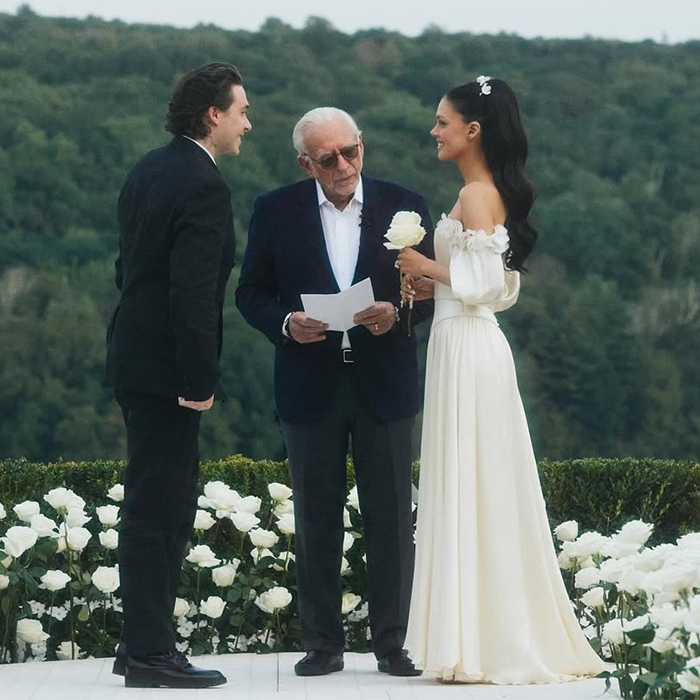 Couple standing with officiant during outdoor wedding ceremony surrounded by white flowers and greenery.