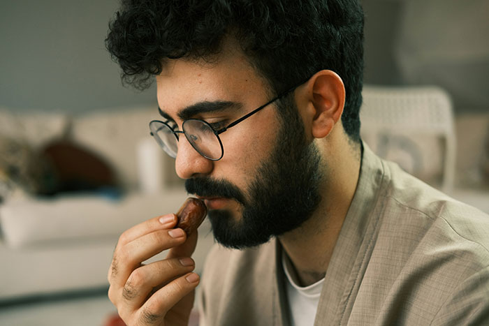 Young man with glasses and beard, deep in thought while holding a small object, reflecting on love and relationships.