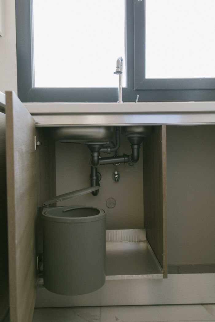 Open kitchen cabinet under sink showing plumbing pipes and attached trash bin in modern home setting.