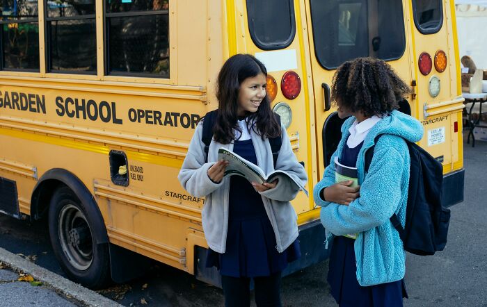 Two schoolgirls talking by a yellow bus, illustrating people ask Americans if these things are true in real life.