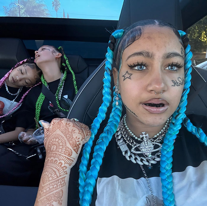 North West with blue braids and facial tattoos sitting in a car, highlighting Kanye West&rsquo;s warning about her online life.