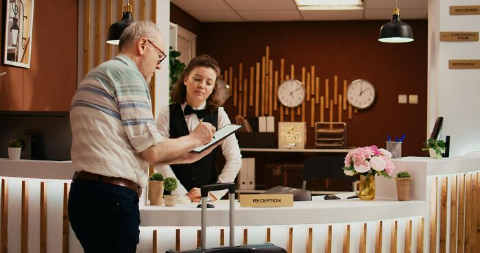 Elderly man misreading social cues while checking in at hotel reception with attentive staff behind the desk.