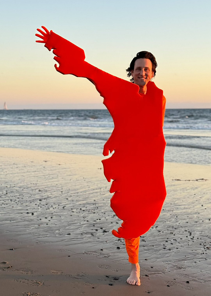One-legged man wearing a vibrant red Halloween costume posing on the beach at sunset, smiling with raised arm.
