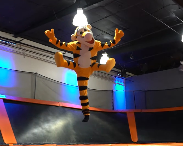 One-legged man jumps on trampoline wearing a colorful Halloween costume in an indoor trampoline park with blue lights.