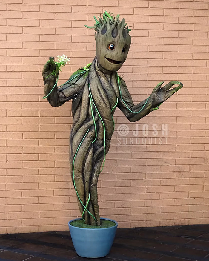 One-legged man in detailed Groot Halloween costume standing in a blue pot against a brick wall background.