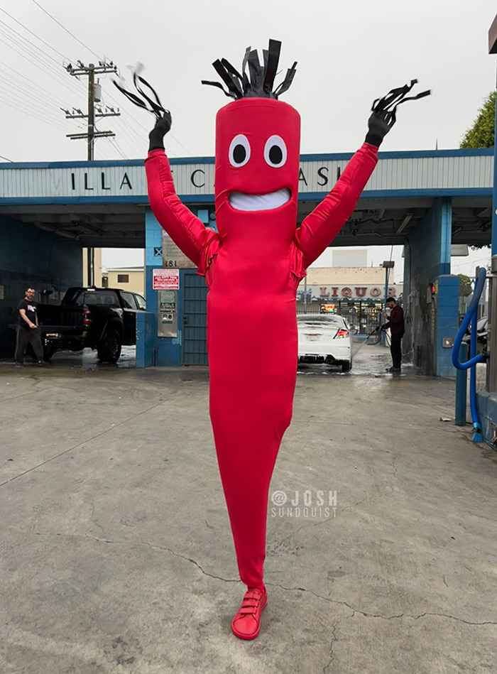 One-legged man in a vibrant red inflatable costume with black gloves and animated arms at an outdoor car wash.